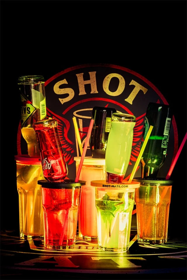 Fünf bunte Gläser mit eingesetzten Flaschen und Shot-Mate Booze Dock Aufsätzen vor beleuchtetem Logo.