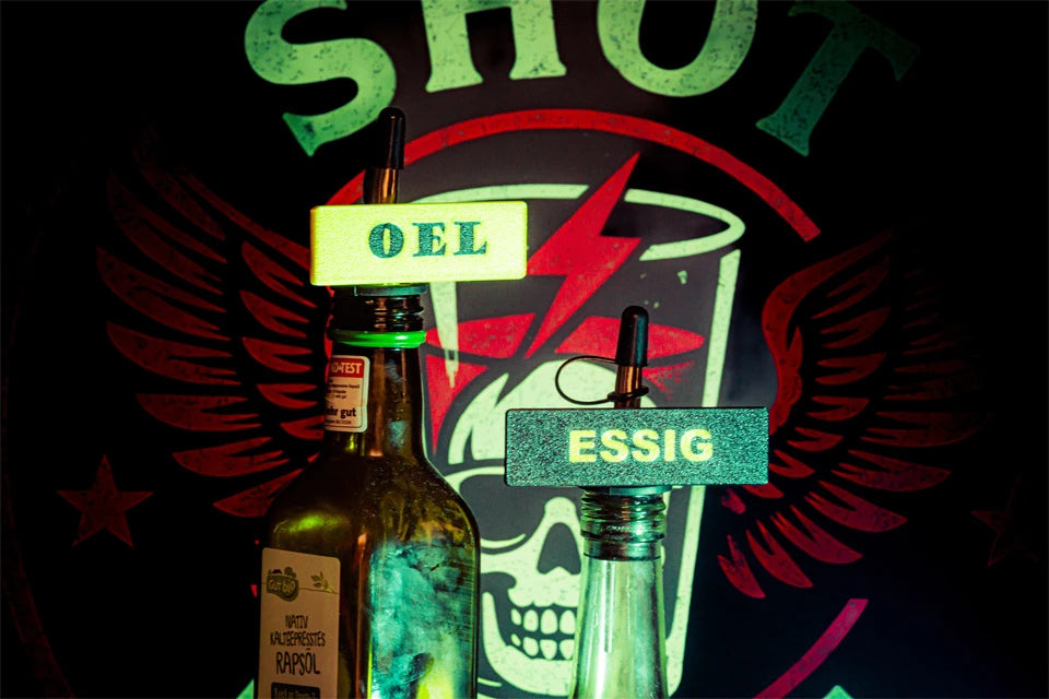Zwei Glasflaschen mit Shot-Mate-Clips beschriftet mit „Öl“ und „Essig“ vor einem leuchtenden Hintergrund.
