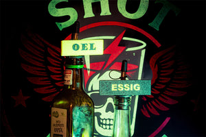 Zwei Glasflaschen mit Shot-Mate-Clips beschriftet mit „Öl“ und „Essig“ vor einem leuchtenden Hintergrund.