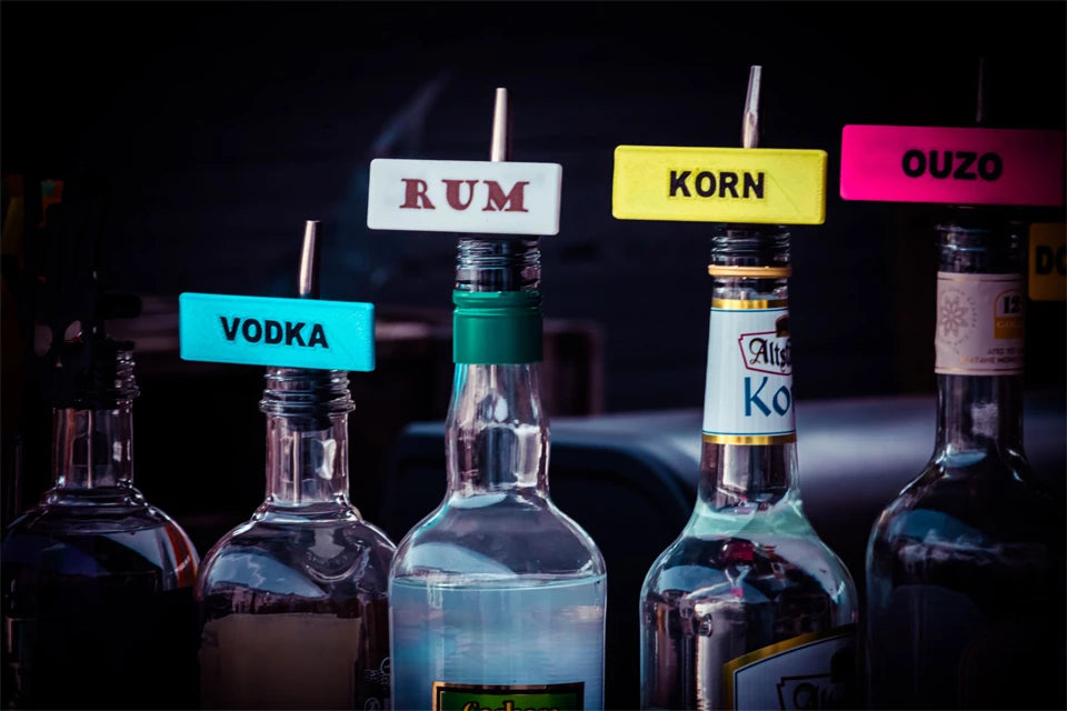 Flaschen mit bunten Shot-Mate-Clips zur Kennzeichnung von Spirituosen wie Vodka, Rum, Korn und Ouzo.