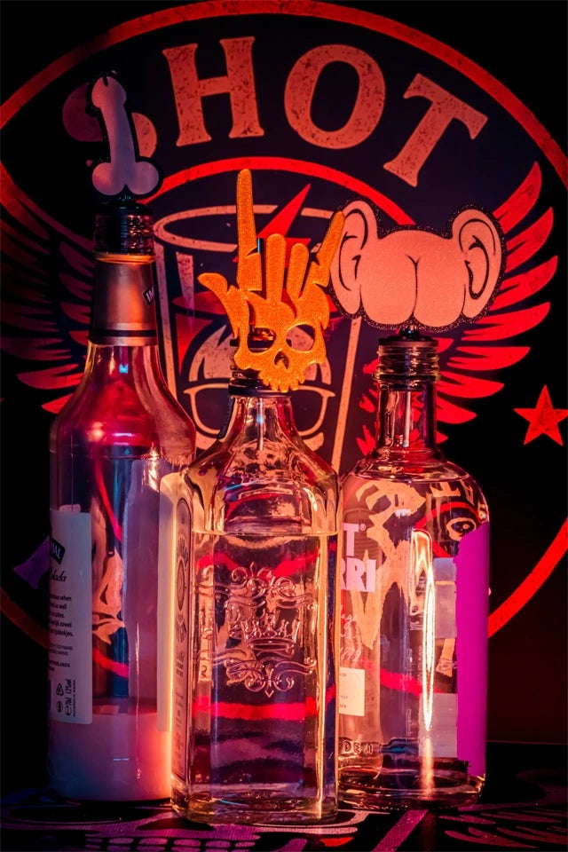 Drei Flaschen mit Shot-Mate-Aufsätzen in Penis-, Rockerhand- und Po-Form vor beleuchtetem Markenlogo.