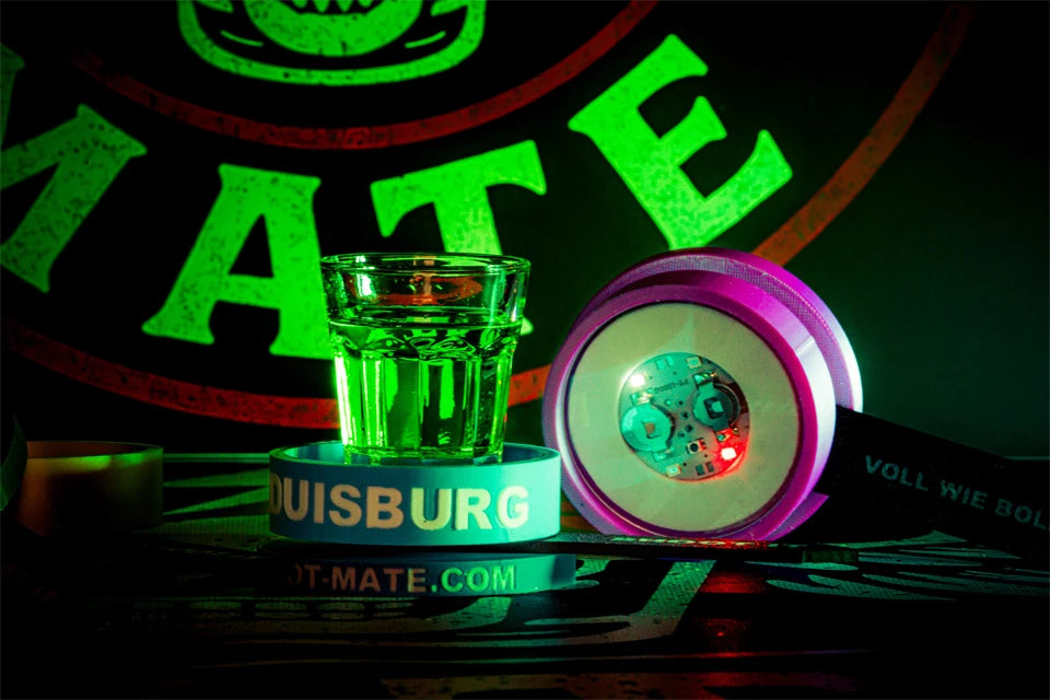Leuchtender LED-Spinner, Shotglas mit grünem Drink und Silikonarmbänder vor Shot-Mate-Logo.
