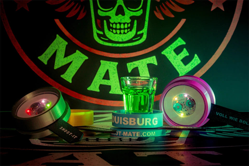 Zwei leuchtende LED-Spinner mit grünem Shotglas und Shot-Mate-Armbändern vor Markenlogo.