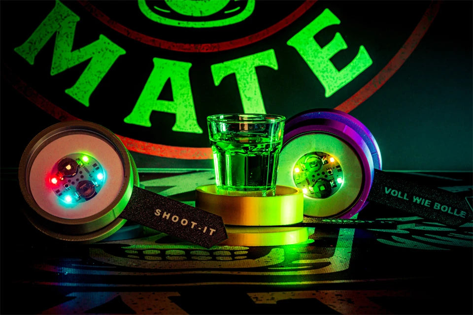 Grünes Shotglas zwischen zwei leuchtenden LED-Spinnern mit Shot-Mate-Armbändern vor Markenlogo.