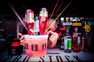 Person zeigt auf den Shot-Mate Booze Pott Getränkespender mit mehreren Cocktailflaschen. Das System eignet sich für Cocktails, Longdrinks, Partys und Veranstaltungen.